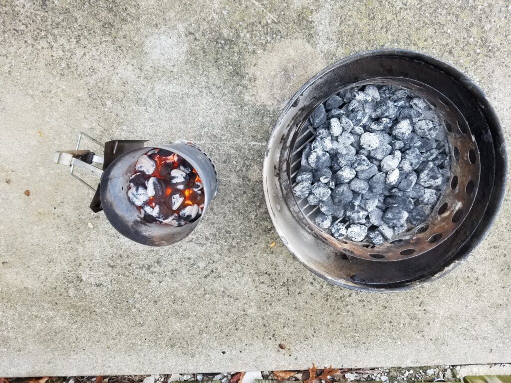 Lit charcoal in a chimney starter next to a charcoal ring of unlit briquettes for the minion method