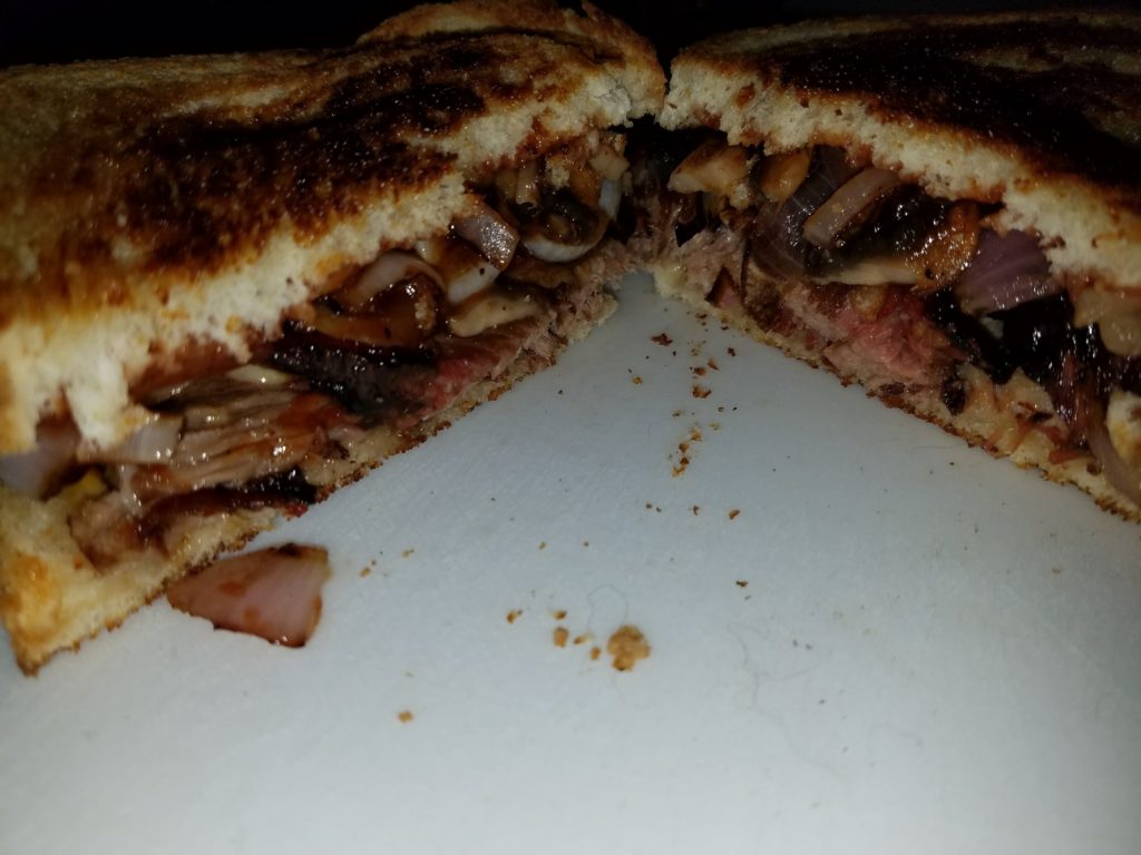 The finished brisket sandwich, cut in half, showing the melted cheese, brisket, and sautéed onions and mushrooms