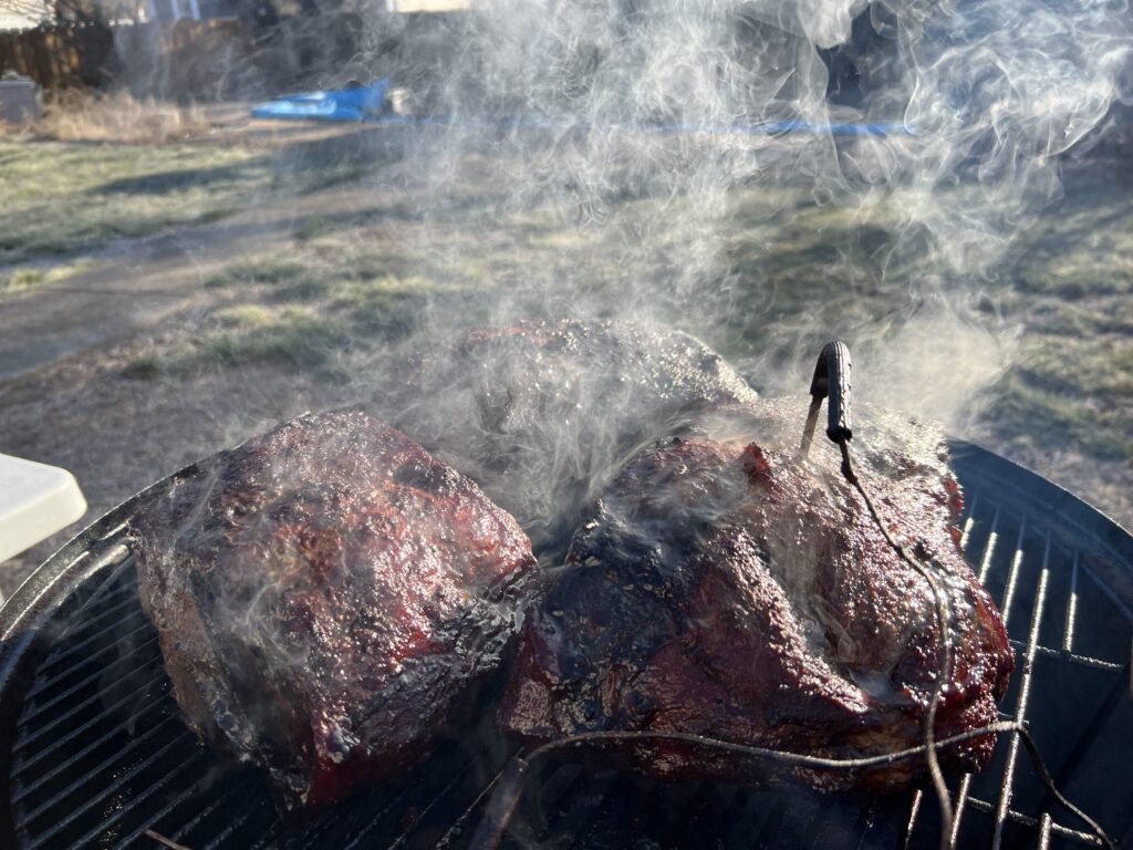 Two pork butts with dark bark on the smoker grate, smoke rising