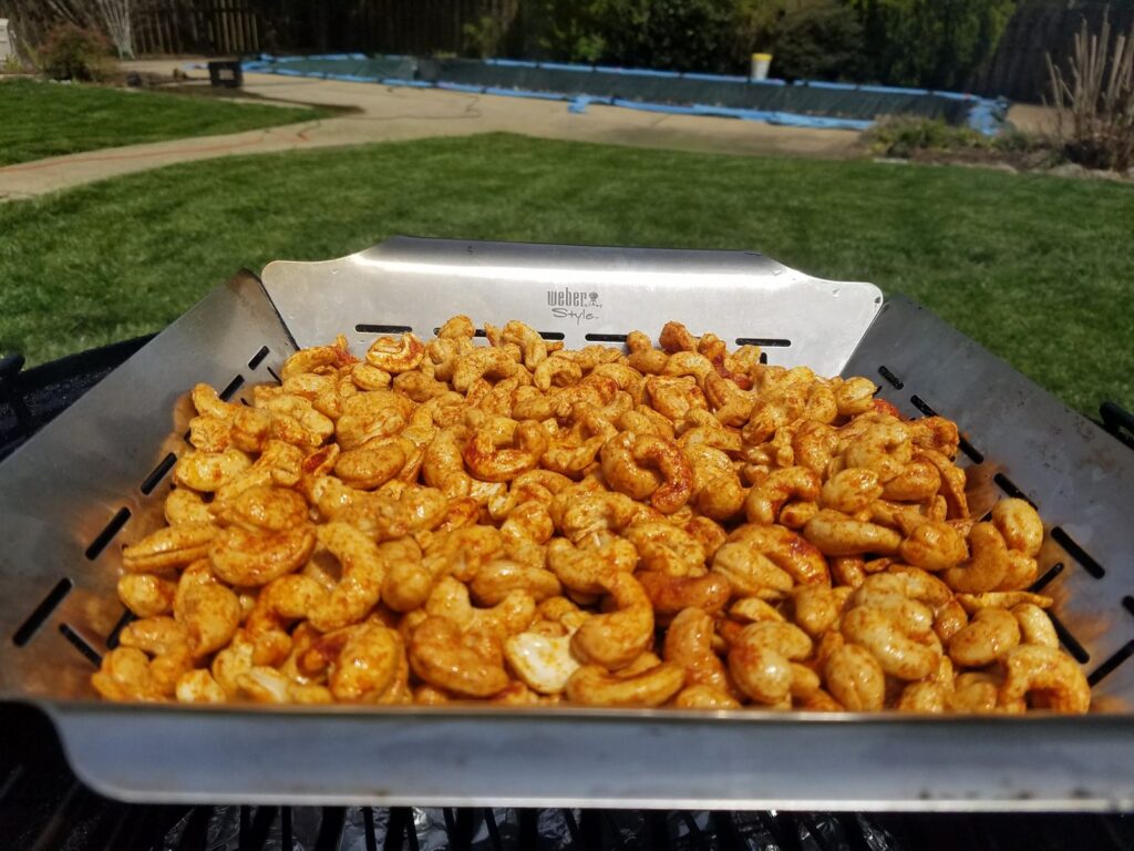 Seasoned cashews in a Weber Style slotted grill pan on a grill at the start of the cook, backyard lawn and covered pool behind